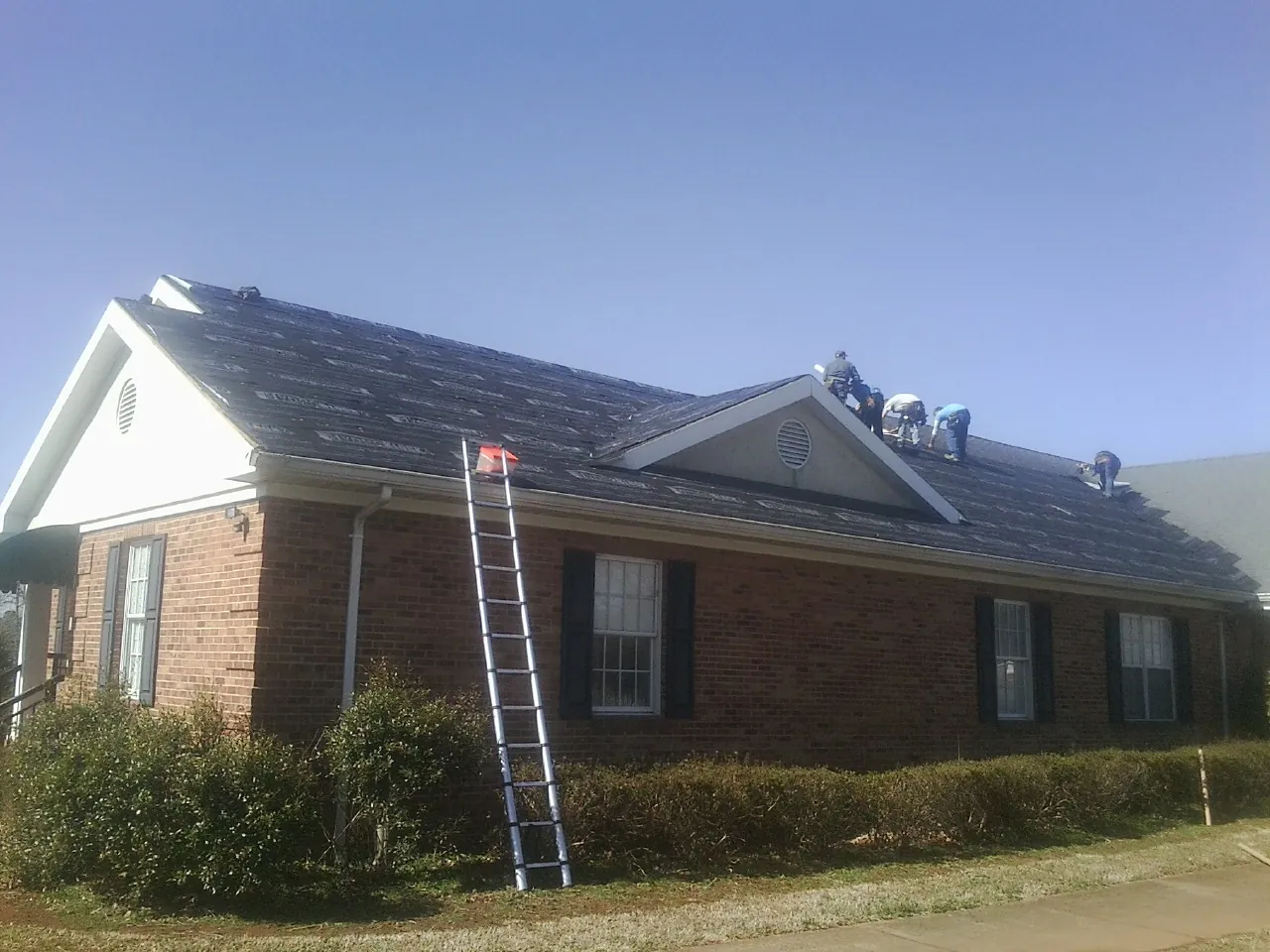 Crew tearing shingles off a brick ranch roof with a ladder set — Emergency Roof Repair work by our crew in Corunna
